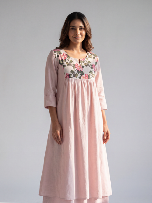 A woman modeling a light pink cotton kurta with a floral embroidered yoke, standing against a neutral gray studio background.