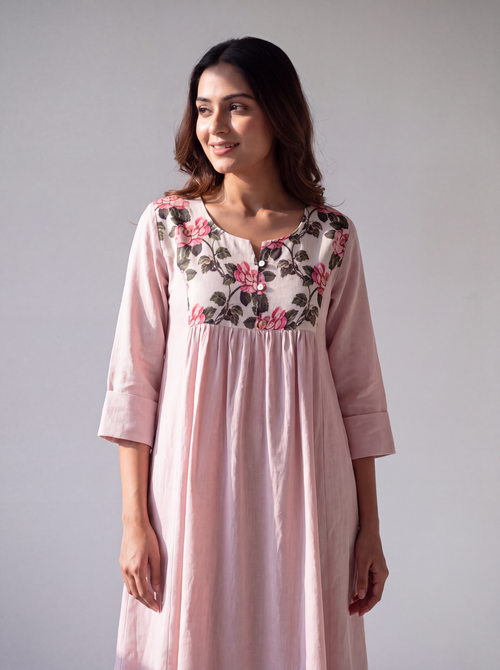 A woman modeling a light pink cotton nighty dress with a floral embroidered yoke in pink, green, and black, set against a plain light gray studio background.