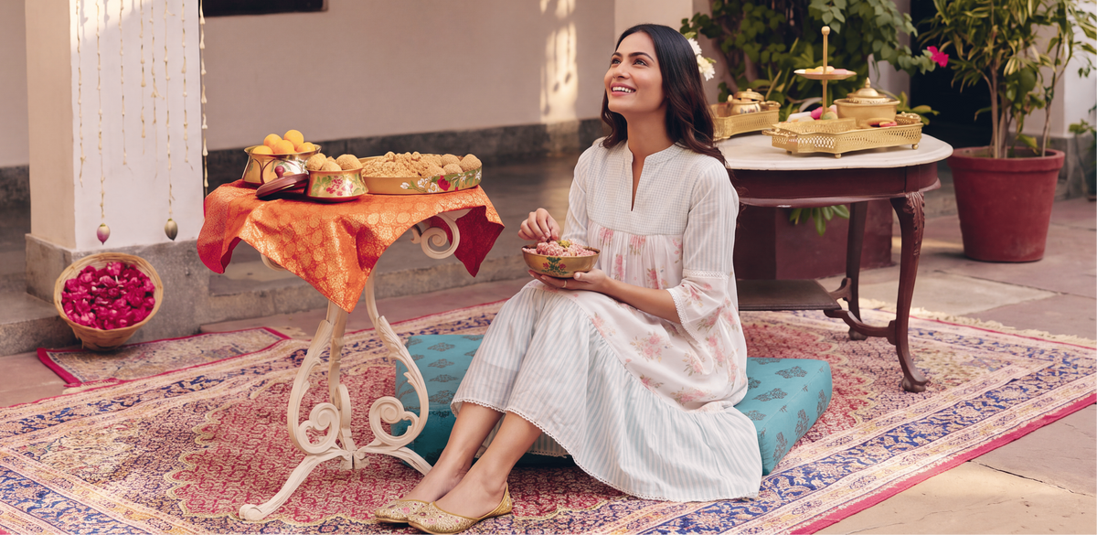 A woman in a white traditional outfit sits on a teal cushion on a patterned rug in an outdoor courtyard, holding a bowl. A small table beside her holds a tray of sweets, with decorative hanging beads, potted plants, and floral arches in the background.